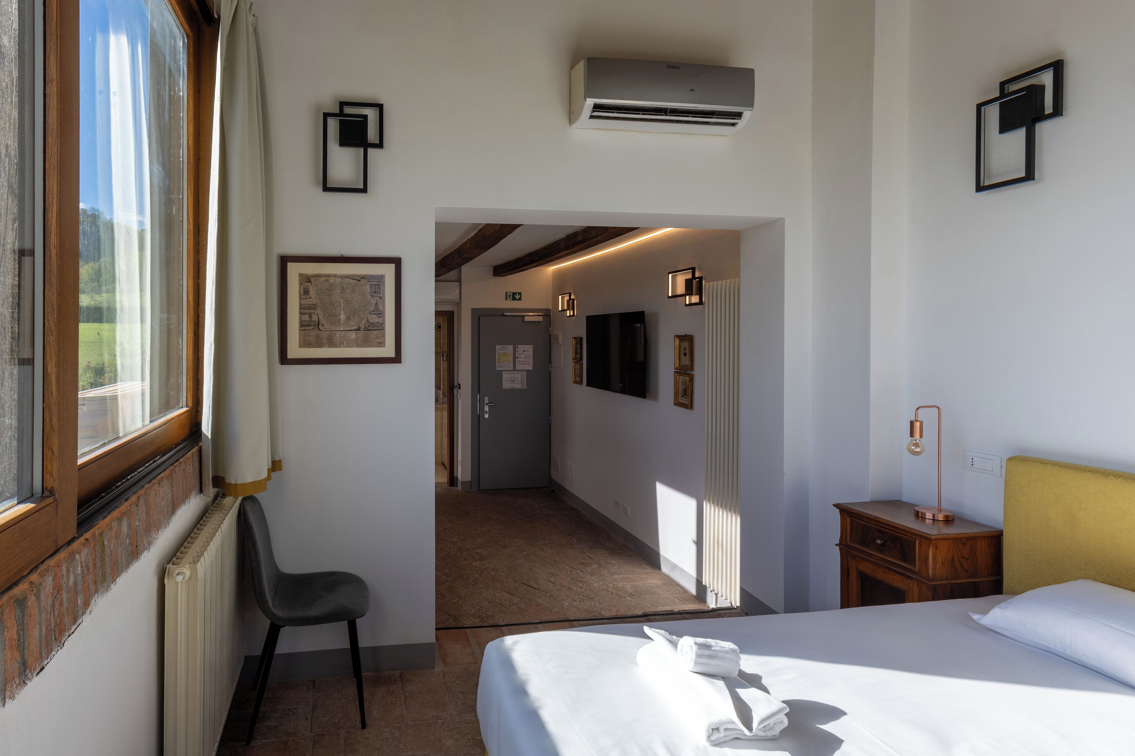 Habitación de hotel con cama, una silla, mesita de noche con lámpara, radiador y ventana con cortinas.