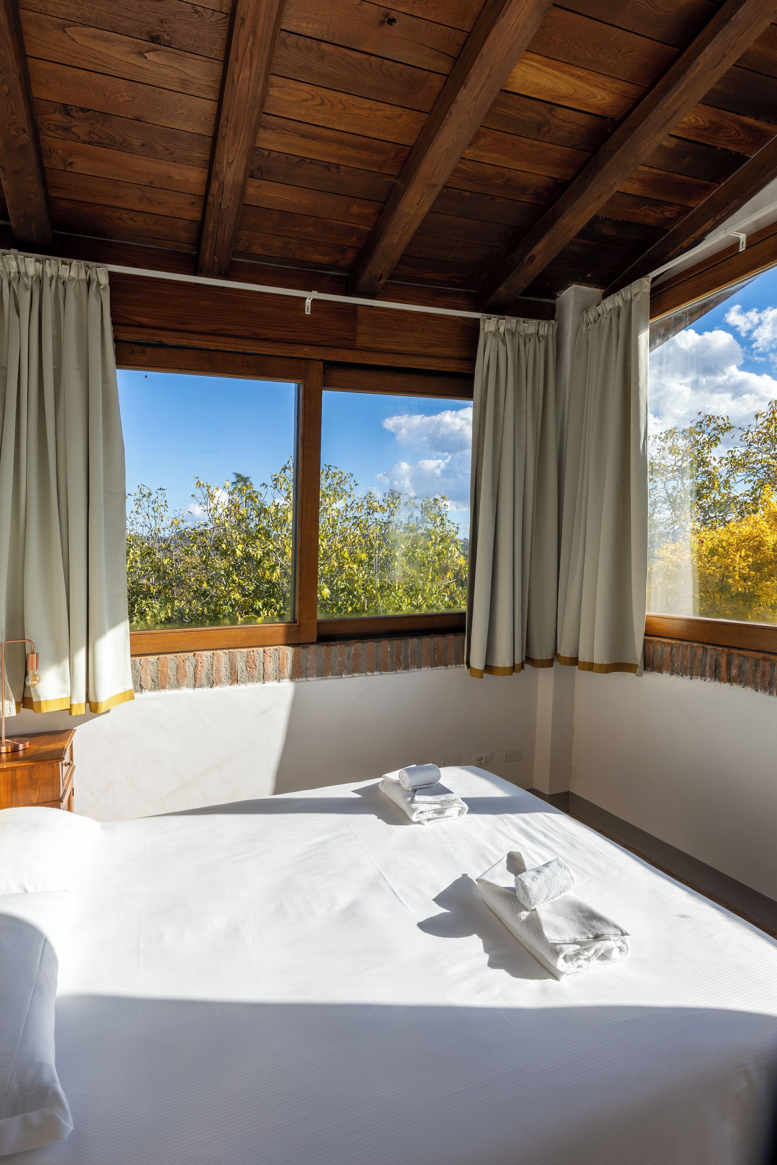 A bedroom with a wooden ceiling, a large bed with white linens, two windows with curtains, and a view of trees.