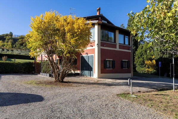 A two-story building with a red and beige facade, a gravel driveway, and a tree with yellow leaves in front. - Borgo Malvasia- La Cascina (Sasso Marconi)