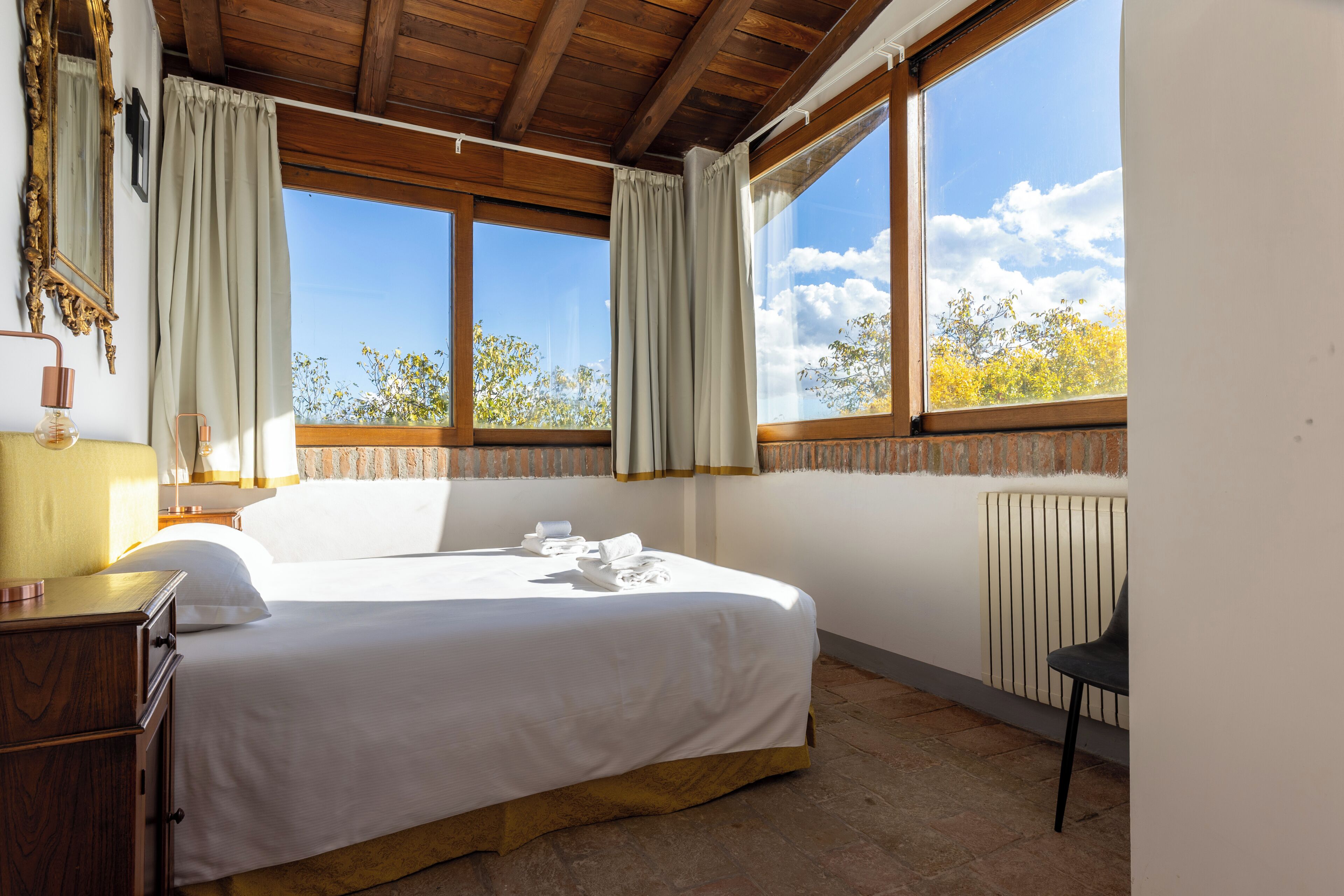 A bedroom with a large bed, wooden ceiling, two windows with curtains, a radiator, and a chair.