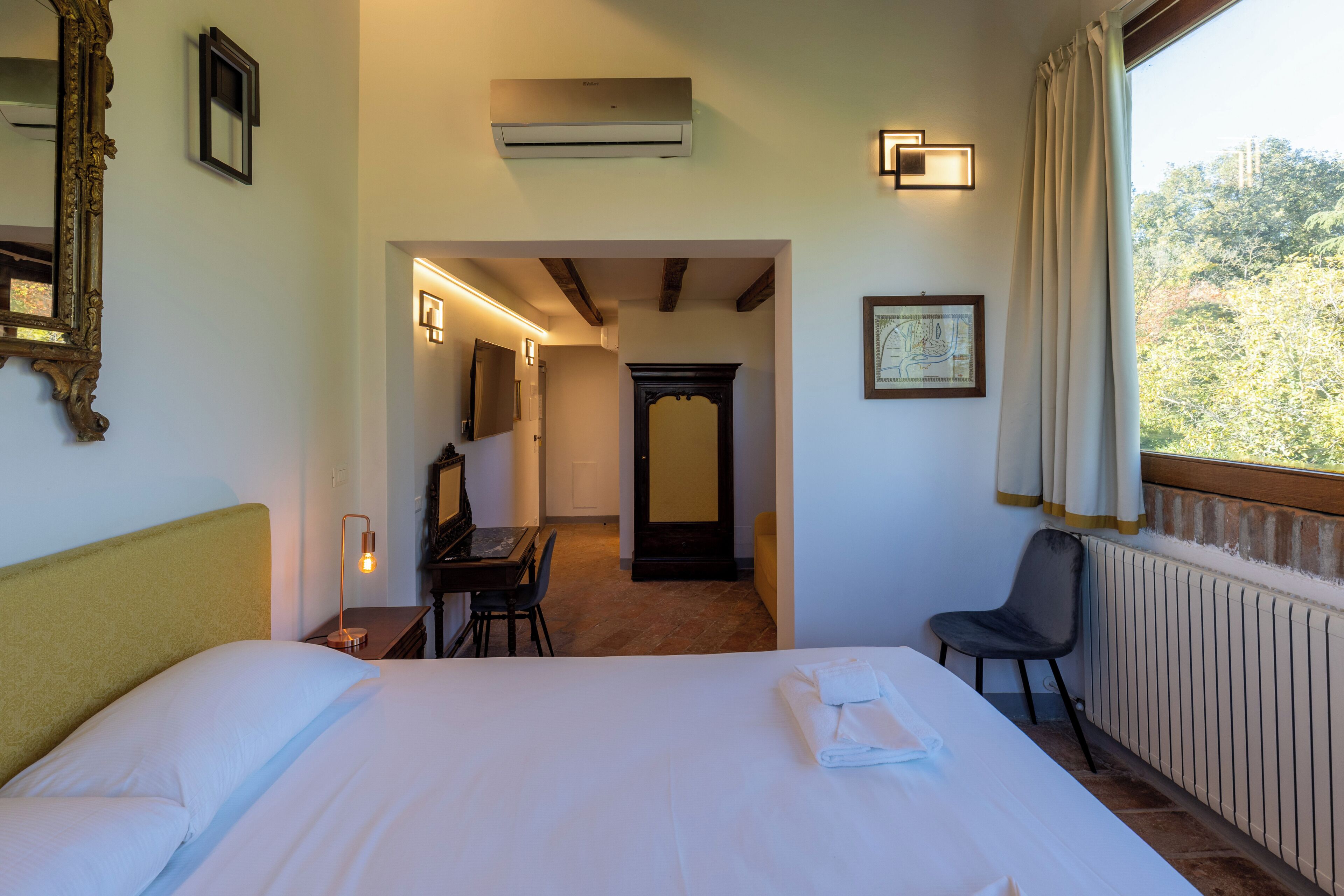 Habitación de hotel con cama, escritorio, silla, espejo y ventana con vista a árboles.