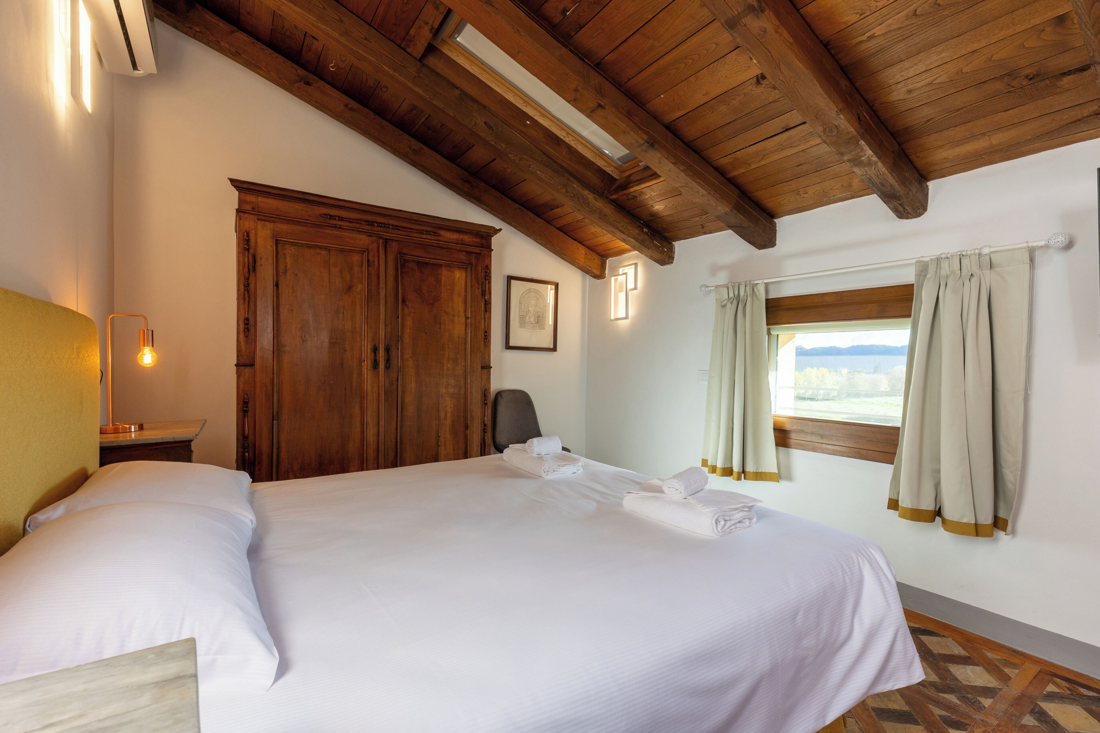 A hotel room with a large bed, wooden ceiling beams, a wardrobe, a desk with a lamp, and a window with curtains.