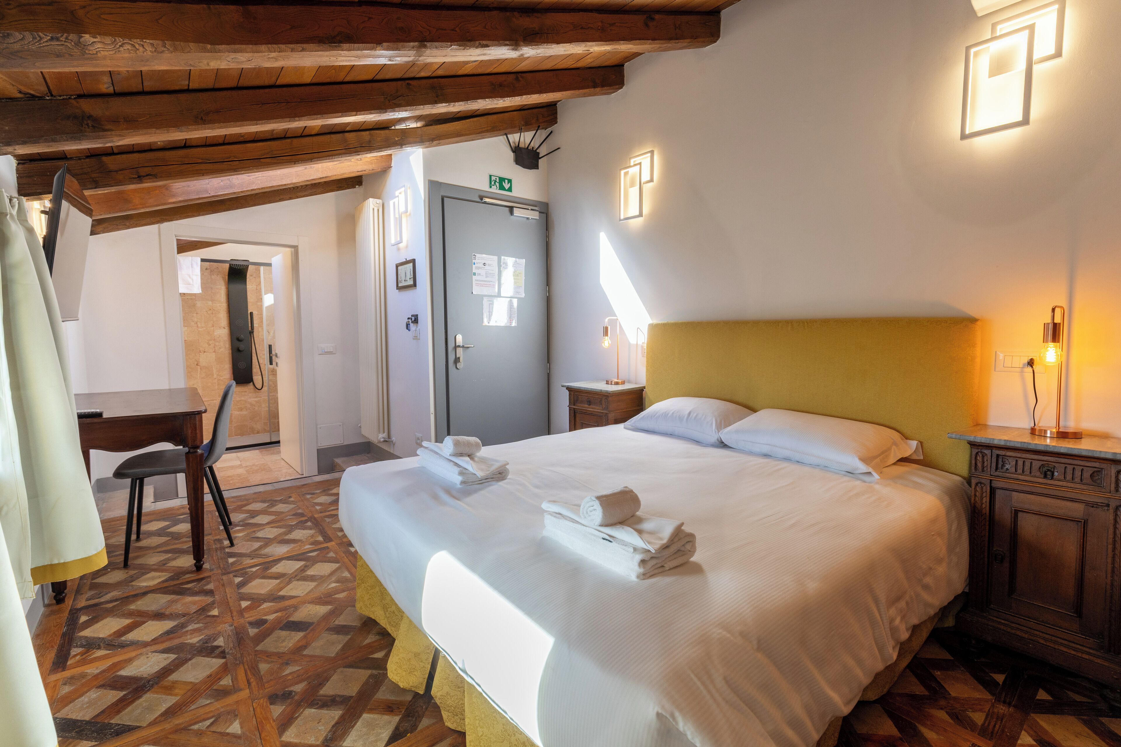 Ein Hotelzimmer mit einem großen Bett, einem hölzernen Kopfteil, einem kleinen Nachttisch mit Lampe, einer Tür mit grünem Fluchtzeichen und einem gemusterten Boden.