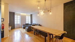 A spacious room with a long wooden table, a bench, and a sofa. There are large windows with curtains, a fire extinguisher, and a blackboard on the wall. - N Apartments - Luís (Porto)