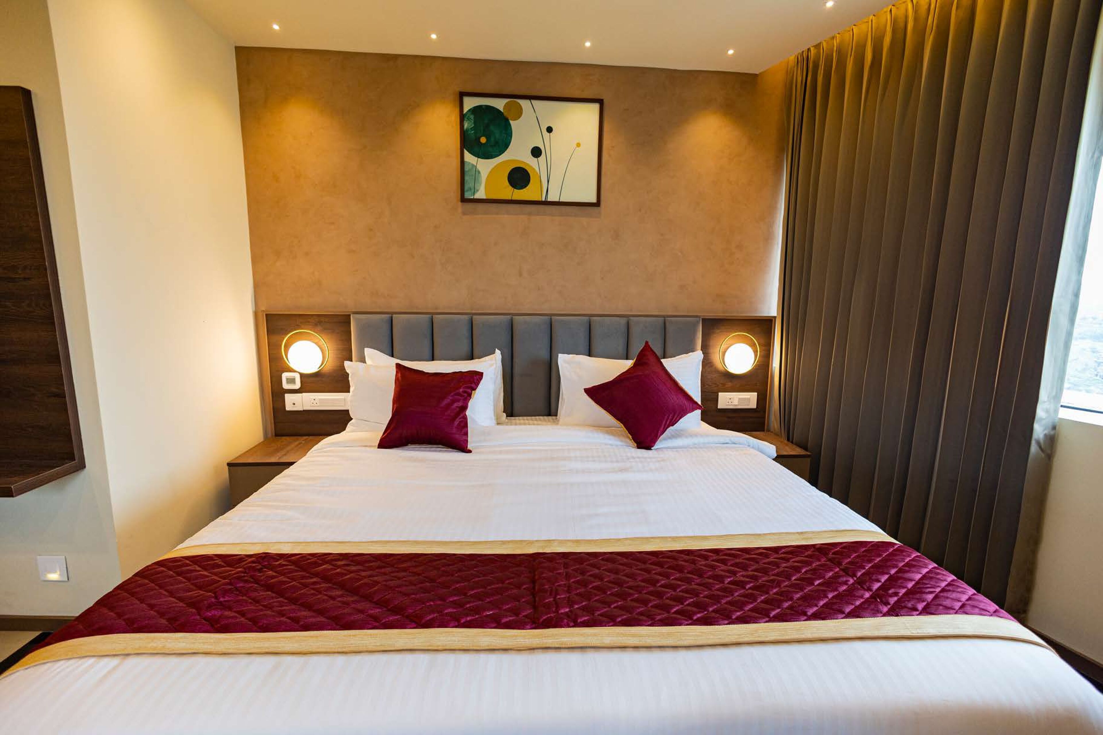 A hotel room with a large bed, two bedside tables with lamps, a headboard with a circular light, and a wall-mounted abstract painting.