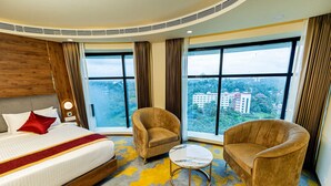 A hotel room with a large bed, two armchairs, a small table, and a view of a cityscape through large windows. - Ascot Hotel Calicut (Kozhikode)