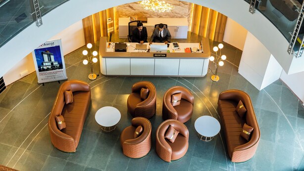 A reception area with a reception desk, two men seated, and four brown leather sofas and two small round tables. - Ascot Hotel Calicut (Kozhikode)