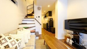 Un salon moderne comprenant un canapé, un téléviseur à écran plat, une table basse en bois et un escalier menant à un espace cuisine.