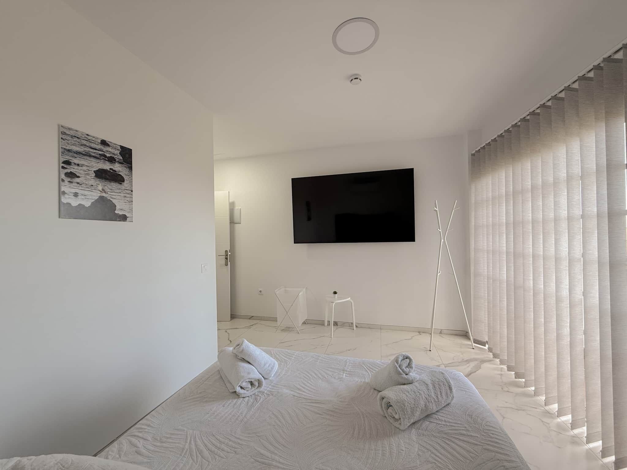 Una camera d'albergo moderna con un letto grande, una TV a schermo piatto fissata al muro e vista sul mare attraverso tende leggere.