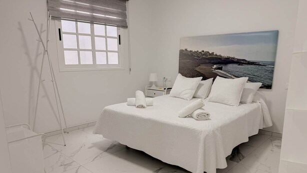 A neatly made bed with white linens and pillows, a painting of a coastal scene on the wall, a window with blinds, and a white floor with marble-like tiles.