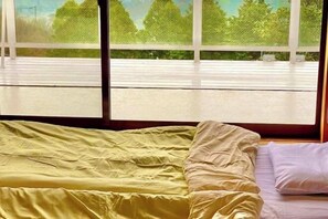 A bed with a yellow bedspread and a white pillow, a wooden floor, and a view of trees through large windows. - Atami Tenku Villa Sky terrace (Atami)