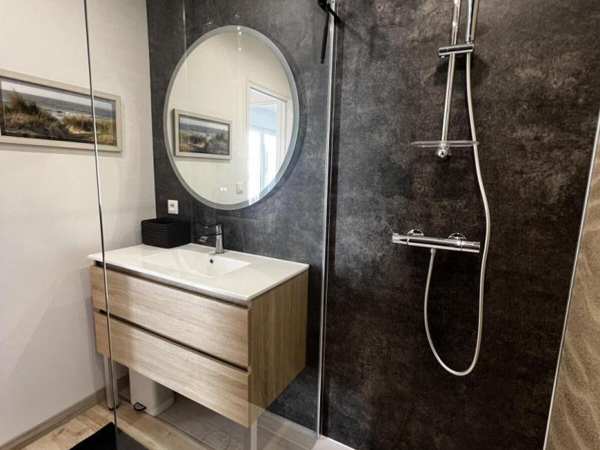 Une salle de bain moderne avec un meuble-lavabo en bois, un miroir rond, une douche équipée d’un pommeau de douche à main et un mur foncé à texture.