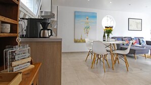 Una cocina moderna con cafetera, un comedor con mesa y sillas blancas, y una sala de estar con sofá y cojines.