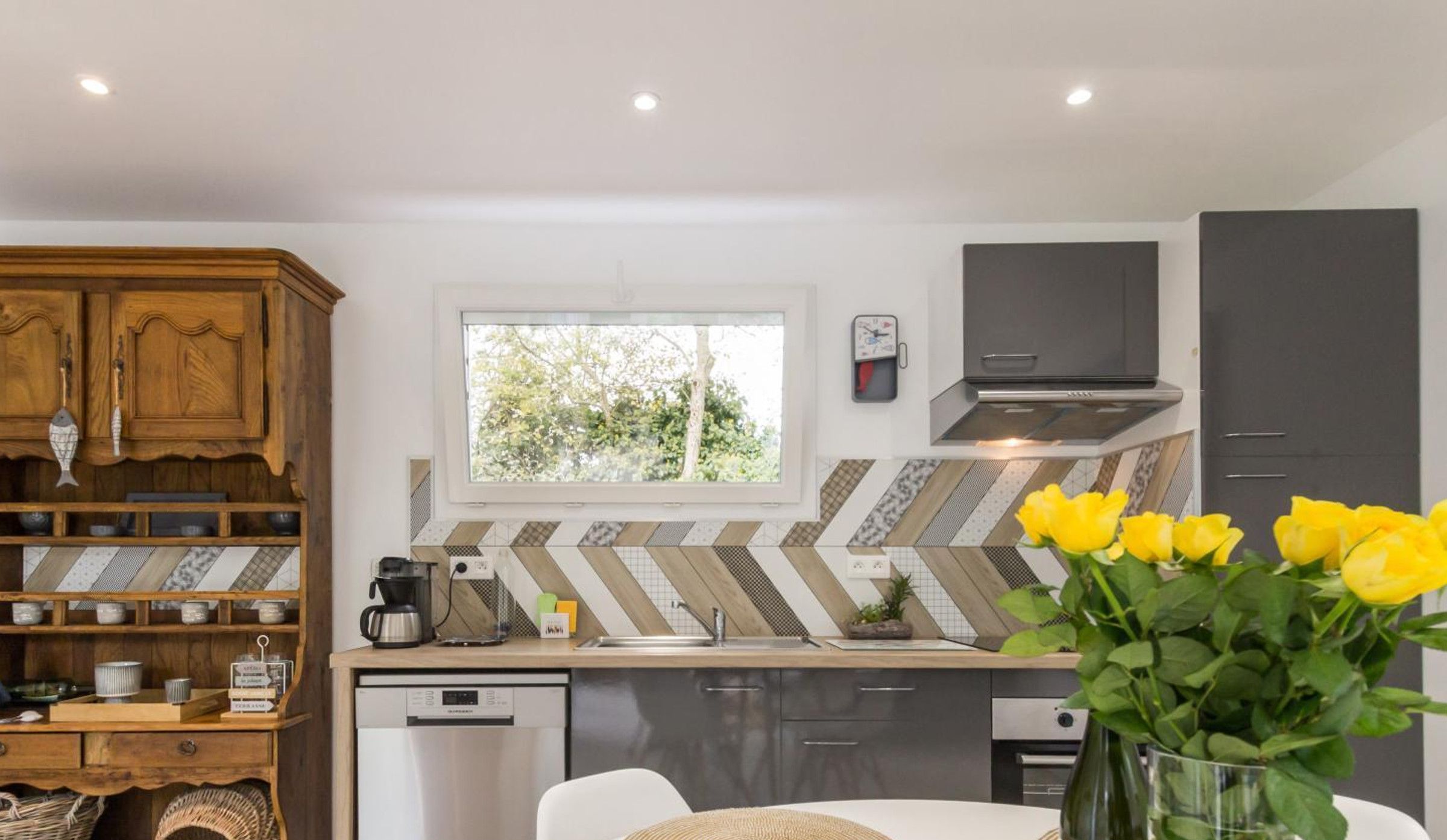 A modern kitchen with a herringbone tile backsplash, a built-in dishwasher, and a window with a view of greenery.