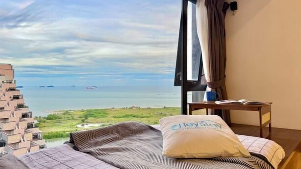 Une chambre avec une grande fenêtre, un lit, une table de chevet avec un livre, et une vue sur la mer.