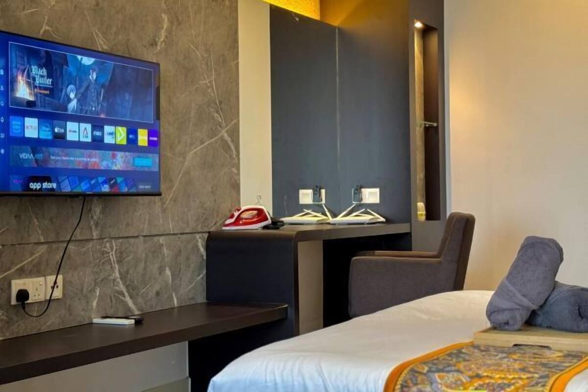 Une chambre d’hôtel moderne dotée d’un grand téléviseur à écran plat fixé au mur, d’un bureau avec un fer à repasser rouge, d’une chaise et d’un lit recouvert d’une courtepointe à motifs.
