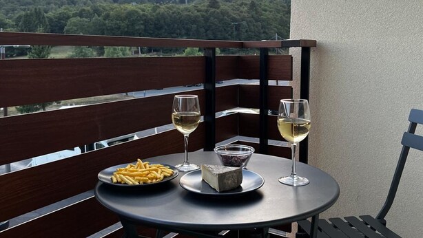 Un balcon avec une table dressée pour deux personnes, où l’on aperçoit une assiette de frites, un plat de fromage et deux verres de vin blanc.