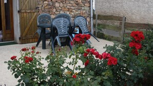 A rustic stone building with a wooden door and a stone wall. There are three black plastic chairs and a table outside. Red flowers are in the foreground. - Country cottage with pets allowed near Roanne (Neulise)