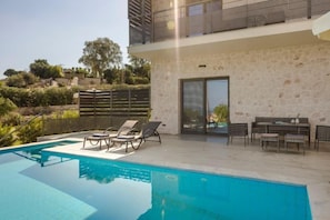 Un espace piscine extérieur moderne, doté de chaises longues et d’une table à manger, entouré d’un mur en pierre et offrant une vue sur le paysage.