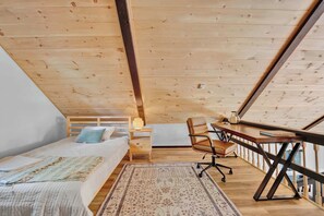 Une chambre avec un plafond en bois, un lit recouvert d’une couverture à motifs, un bureau avec une chaise et une petite table de chevet avec une lampe.