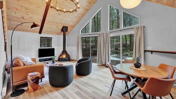 A modern living room with a fireplace, wooden ceiling, large windows, and a dining area. - The Black Pocono Chalet |  Modern Cabin in Nature (Tobyhanna Township)