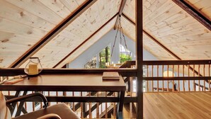 A wooden interior with a sloped ceiling, a table with a candle and book, and a view through a window. - The Black Pocono Chalet |  Modern Cabin in Nature (Tobyhanna Township)