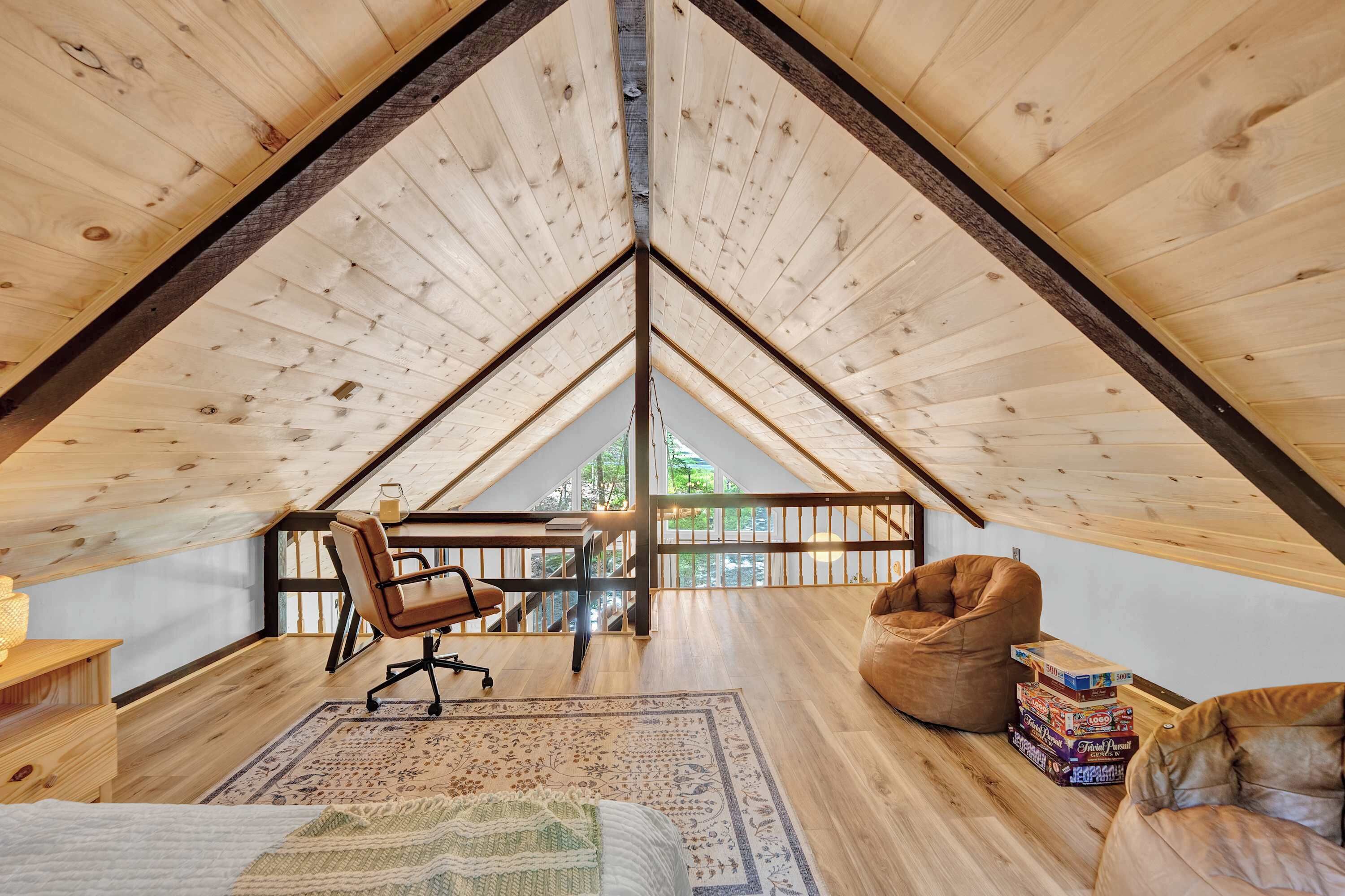 A loft bedroom with a sloped ceiling, a wooden floor, a desk with a chair, a bean bag, and a view of the outdoors.