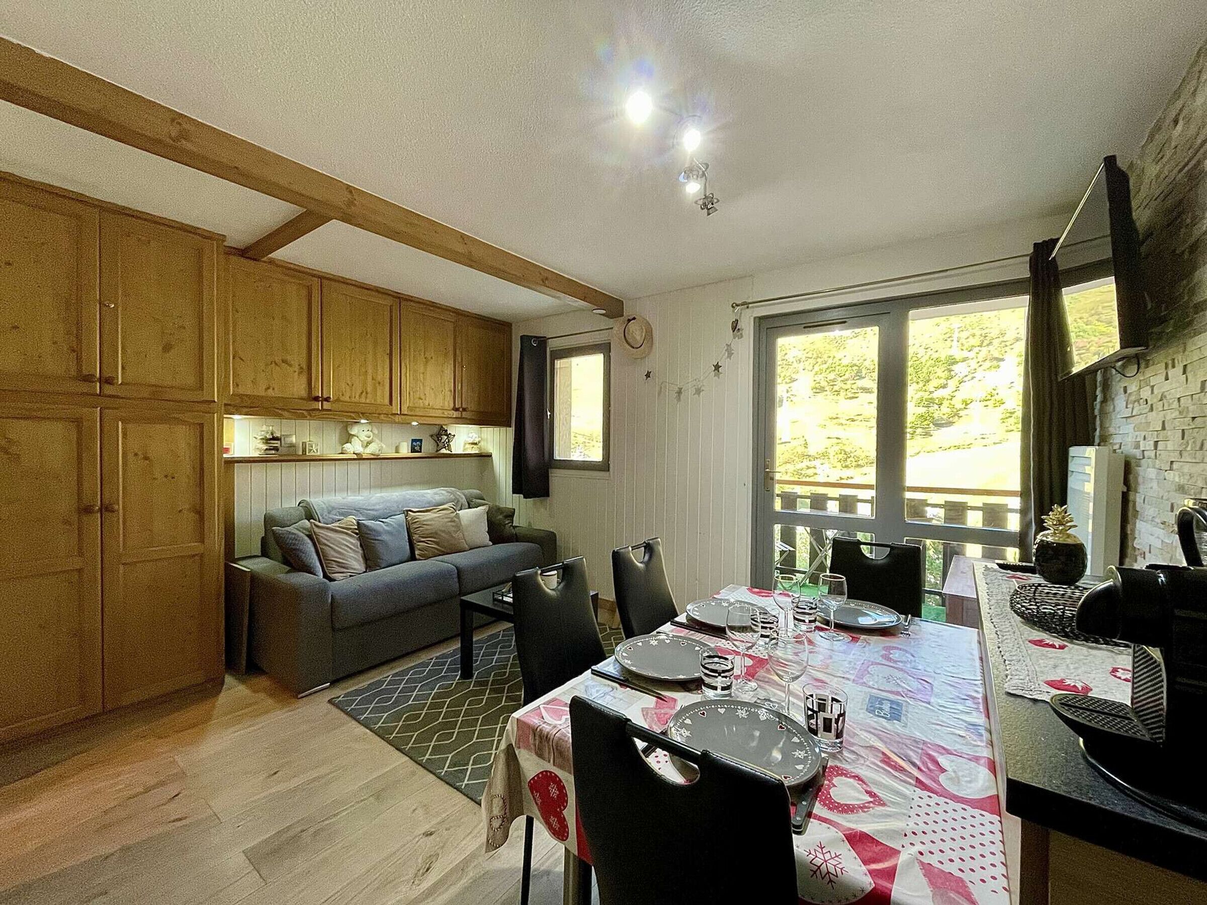 Een gezellige woonkamer met een bank, een eettafel gedekt voor vier personen, houten kasten en een groot raam met uitzicht op groen.