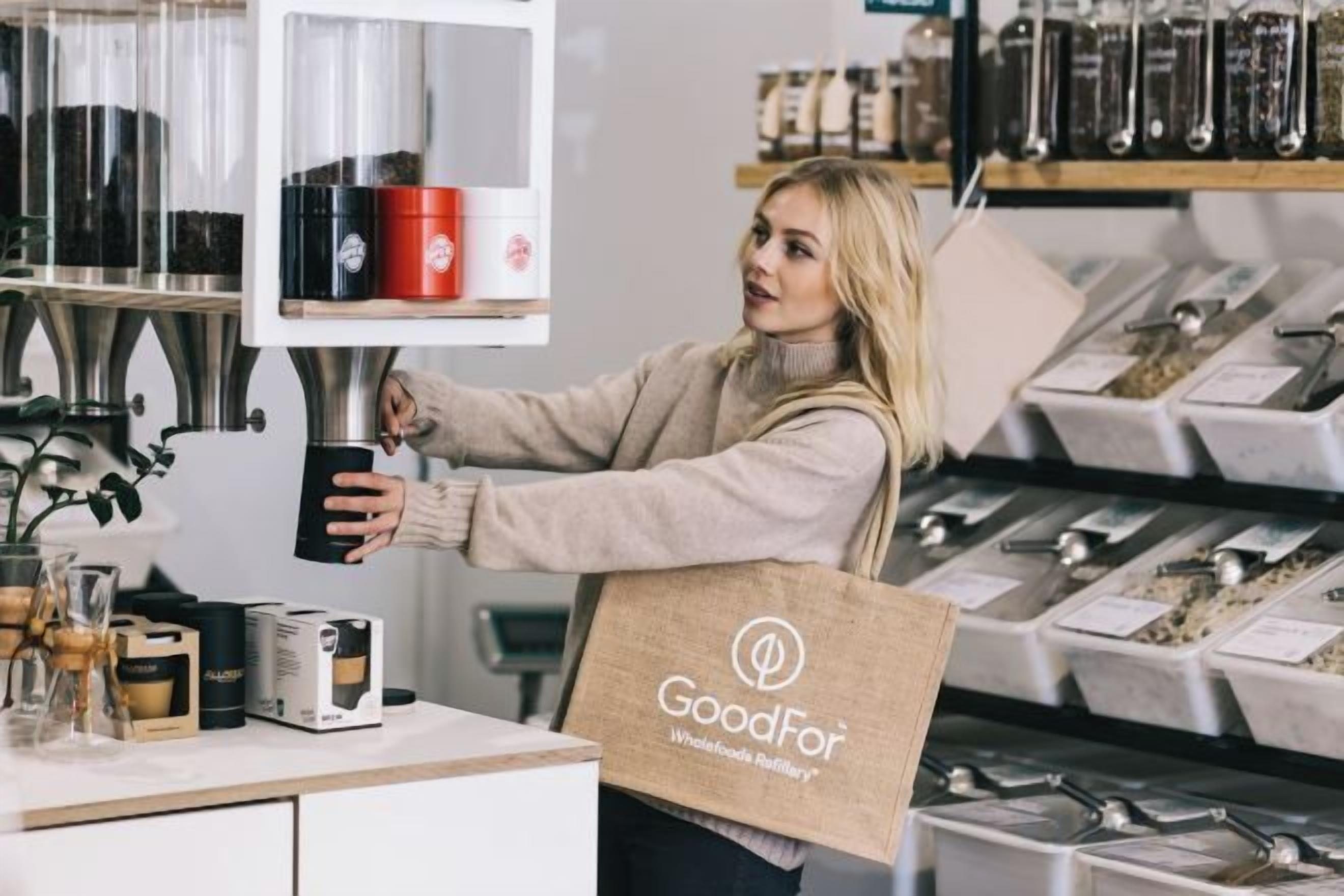 Uma pessoa em uma cozinha com uma máquina de café, prateleiras com recipientes e uma bolsa da marca GoodFor.