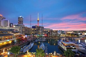A waterfront cityscape with a prominent tower, boats docked along the pier, and buildings with illuminated signs. - Point Residence (Auckland)
