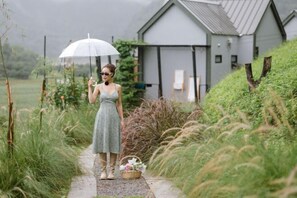 A woman in a dress standing on a path with a basket of flowers, holding an umbrella. - Pause pausa (Phrao)