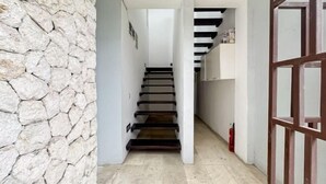 Un couloir étroit, avec un mur de pierre d’un côté et une porte en bois de l’autre. Un escalier mène à l’étage supérieur.
