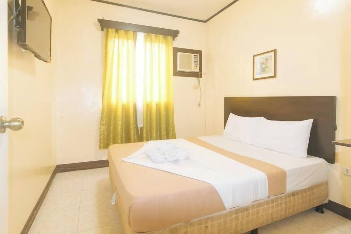 Une chambre d’hôtel avec un lit, des rideaux, un appareil de climatisation, un tableau encadré et un téléviseur fixé au mur.