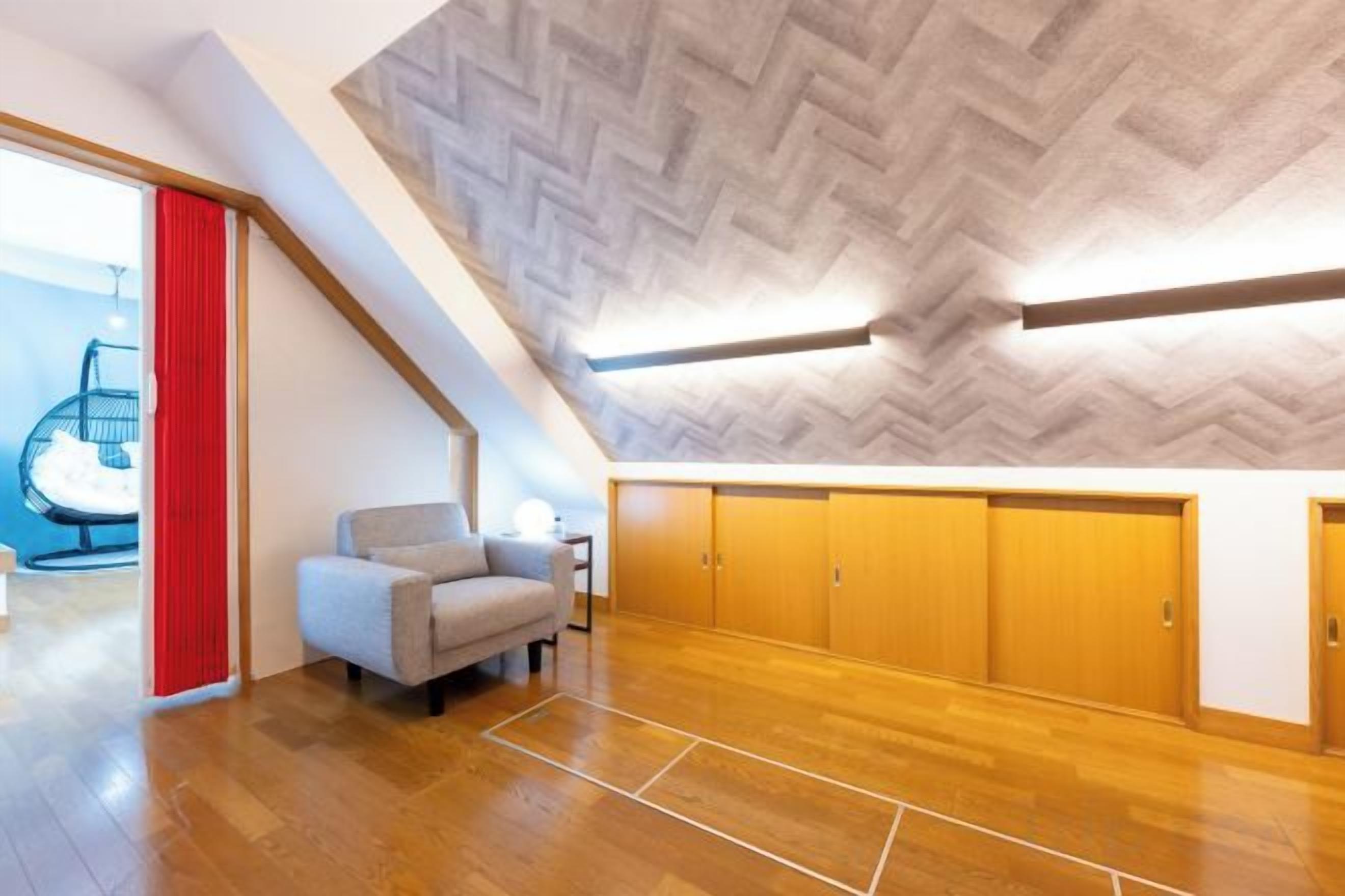 A spacious room with a herringbone-patterned ceiling, wooden flooring, a grey armchair, and a red curtain.