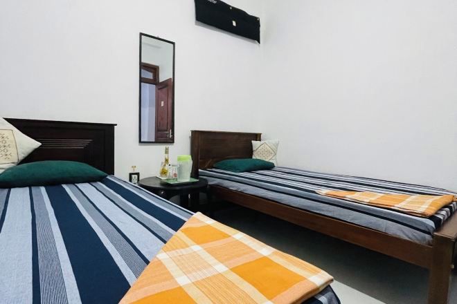 A room with two single beds, each with a different patterned bedspread. There is a small table with a lamp and a bottle on it, and a mirror on the wall.