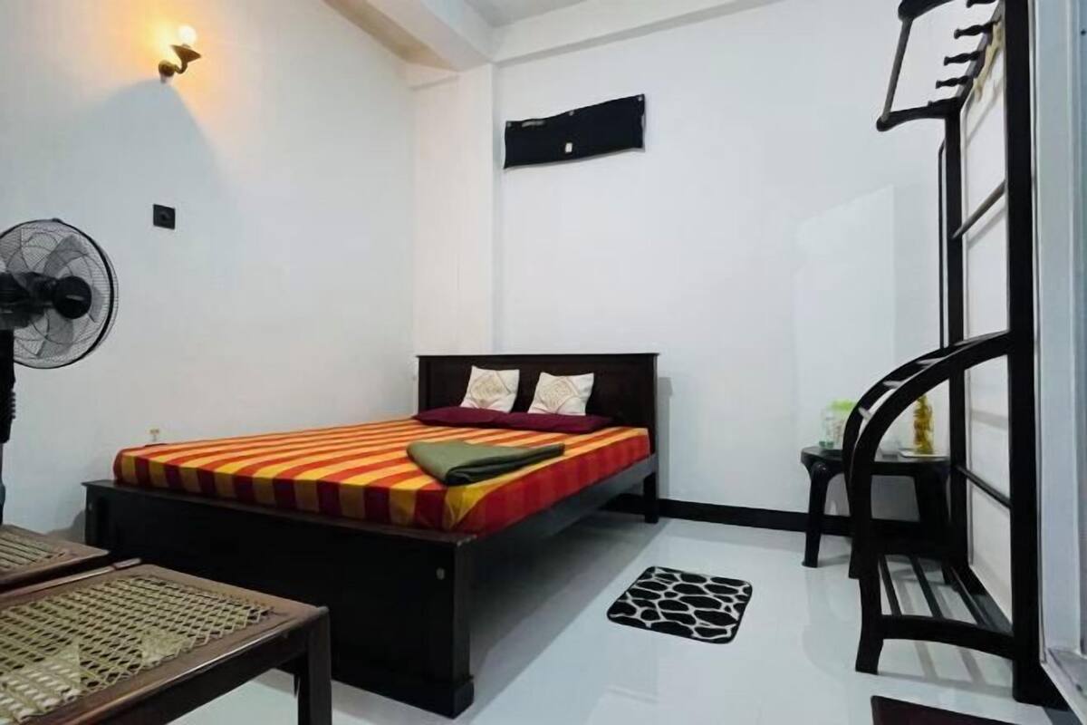 Une chambre à coucher comprenant un lit, un ventilateur, une petite table, un banc et une étagère fixée au mur.