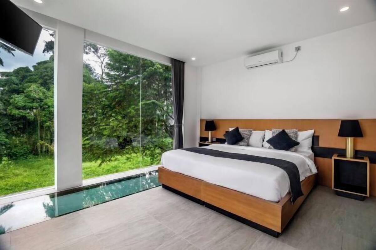 A modern bedroom with a large bed, bedside tables, a TV, and a view of a lush garden.