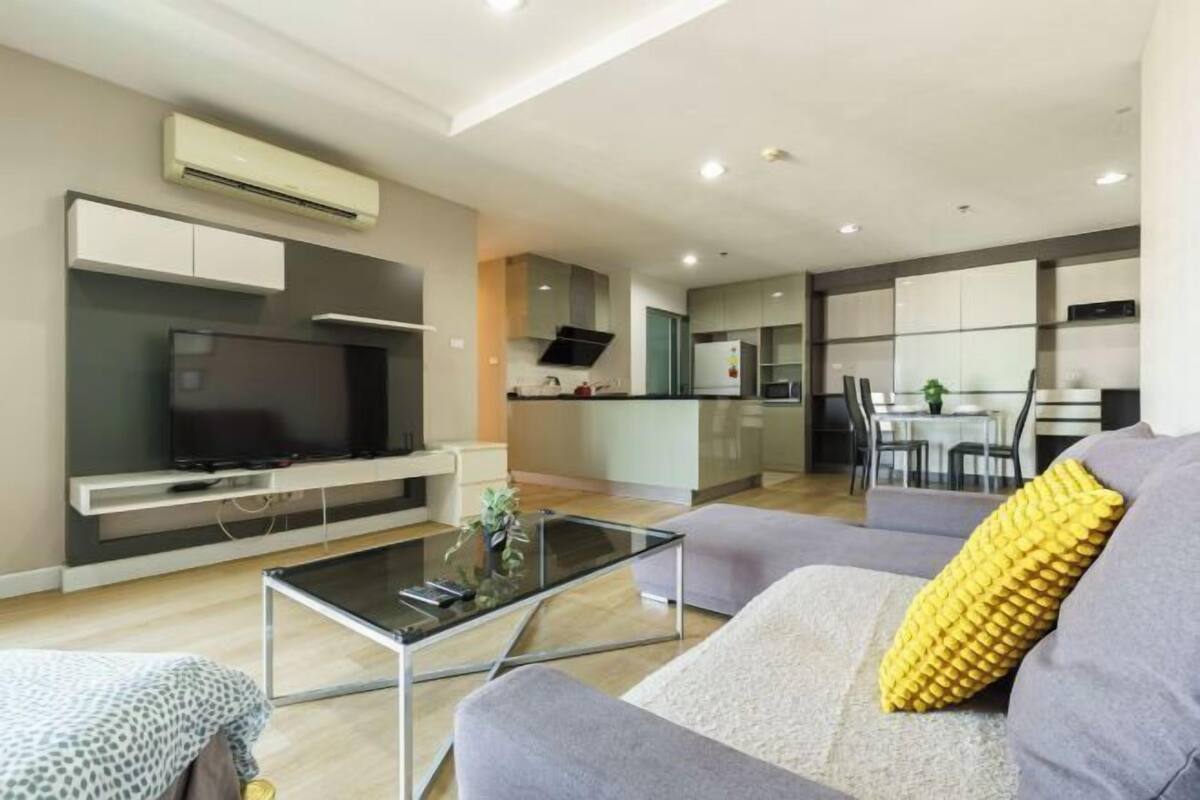 Uma sala de estar moderna com uma televisão de tela plana, uma mesa de centro de vidro e um sofá com um travesseiro amarelo.