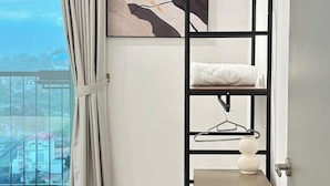 Een kamer met een stapelbed, een witte muur, een abstract schilderij in een lijst, een witte vaas en uitzicht op een stadsgezicht door een raam met gordijnen.