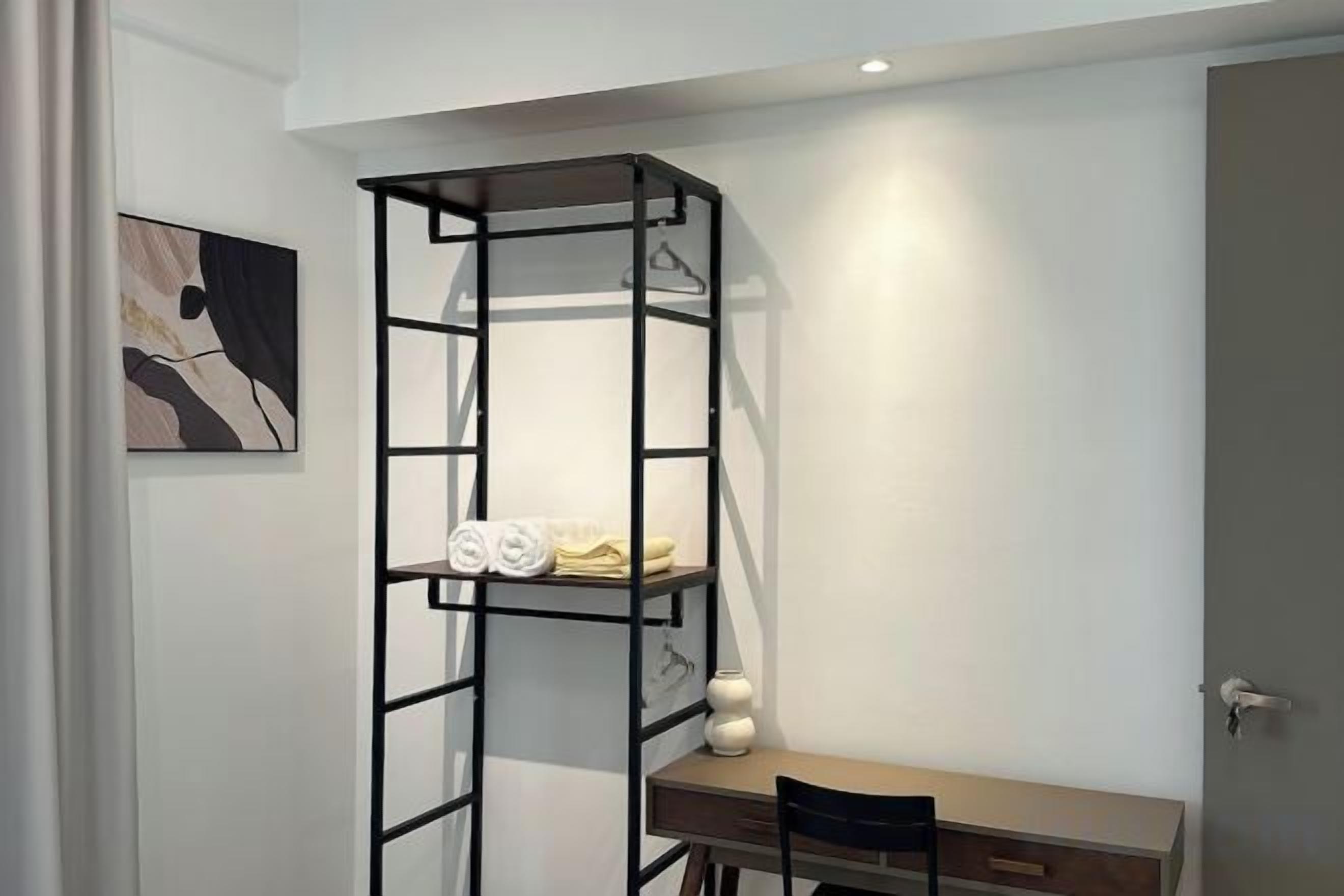 Quarto com escrivaninha, cadeira e uma estante de metal preta encostada a uma parede branca.