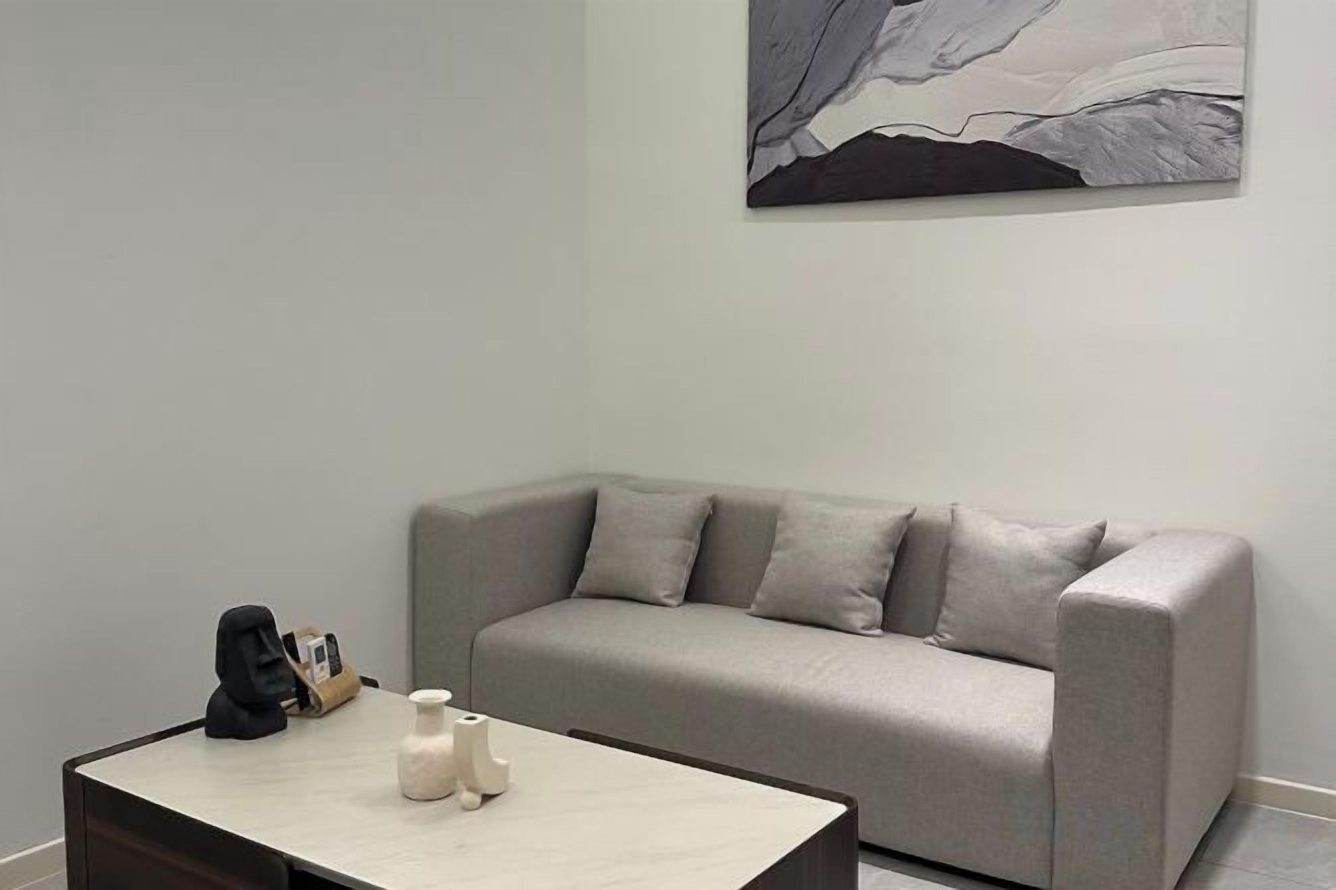 Uma sala de estar moderna com um sofá cinzento, uma mesinha de centro com um vaso e um livro, e um quadro na parede.
