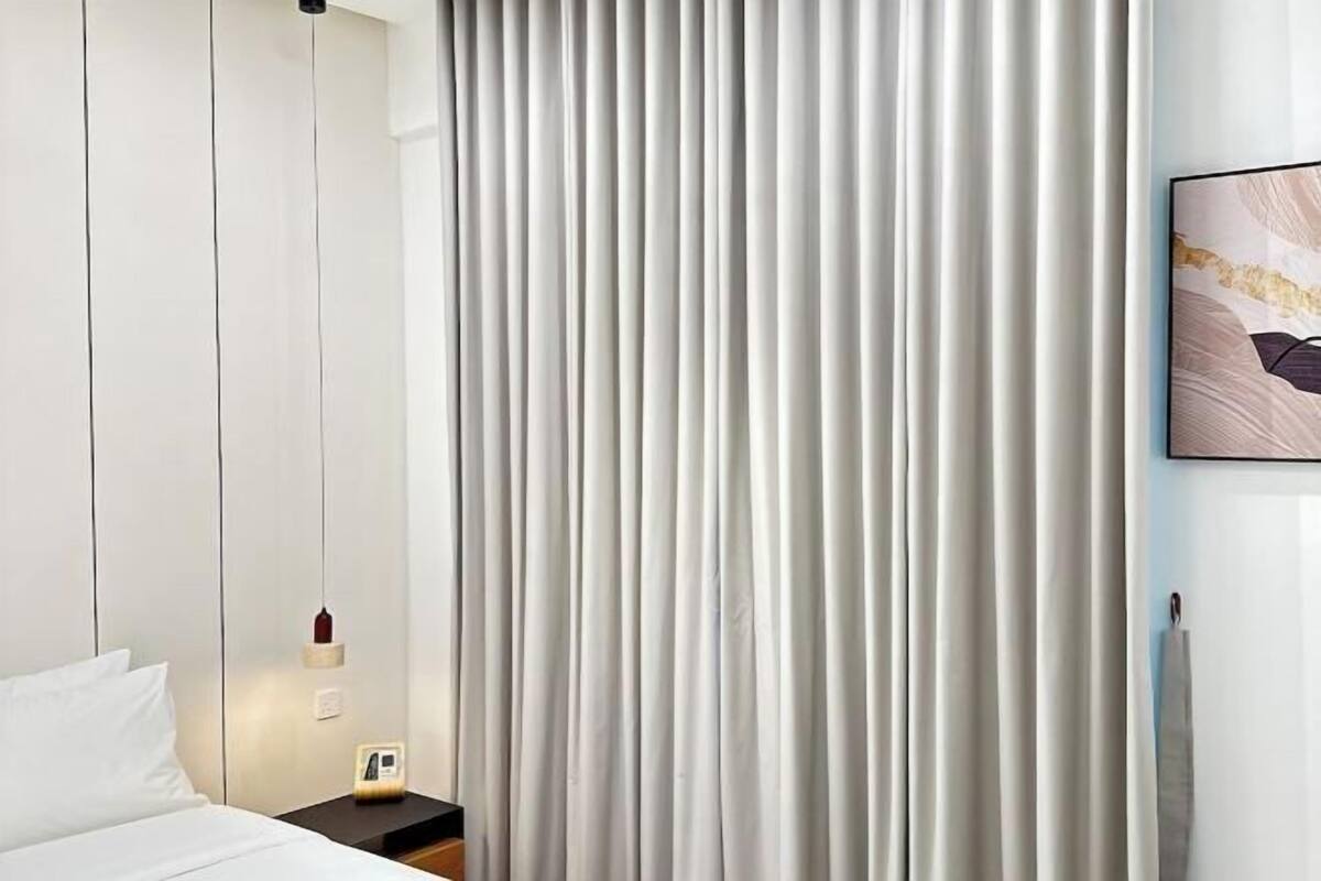 A hotel room with a bed, a nightstand with a lamp, and a large window with vertical blinds.