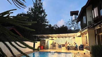 A swimming pool with a covered patio area, surrounded by trees and a building with a water tank on the roof.