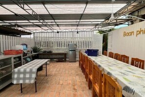 A covered outdoor dining area with a long table, chairs, and a refrigerator.