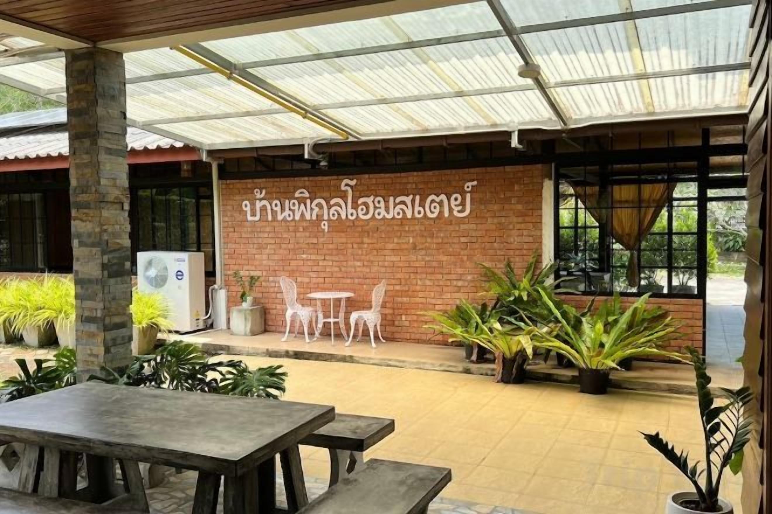 An outdoor patio area with a covered seating area, potted plants, and a brick wall with Thai text.