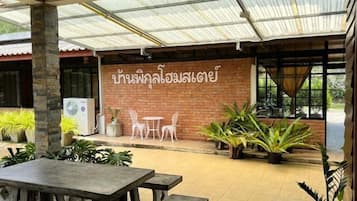 An outdoor patio area with a covered seating area, potted plants, and a brick wall with Thai text.