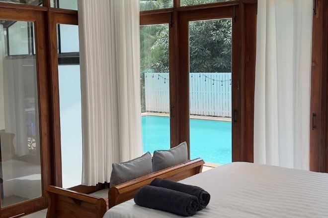 A bedroom with a large window, a bed, a wooden headboard, and a view of a swimming pool.