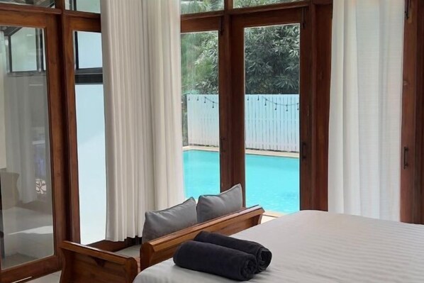 Ein Schlafzimmer mit großem Fenster, einem Bett, einem hölzernen Kopfteil und Blick auf einen Swimmingpool.