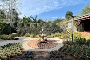 Un jardin avec une fontaine centrale, entouré de sentiers en briques et agrémenté de différentes plantes.