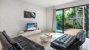 Een moderne woonkamer met een flatscreen televisie, twee leren banken, een salontafel en een grote schuifglazen deur die leidt naar een balkon met planten.